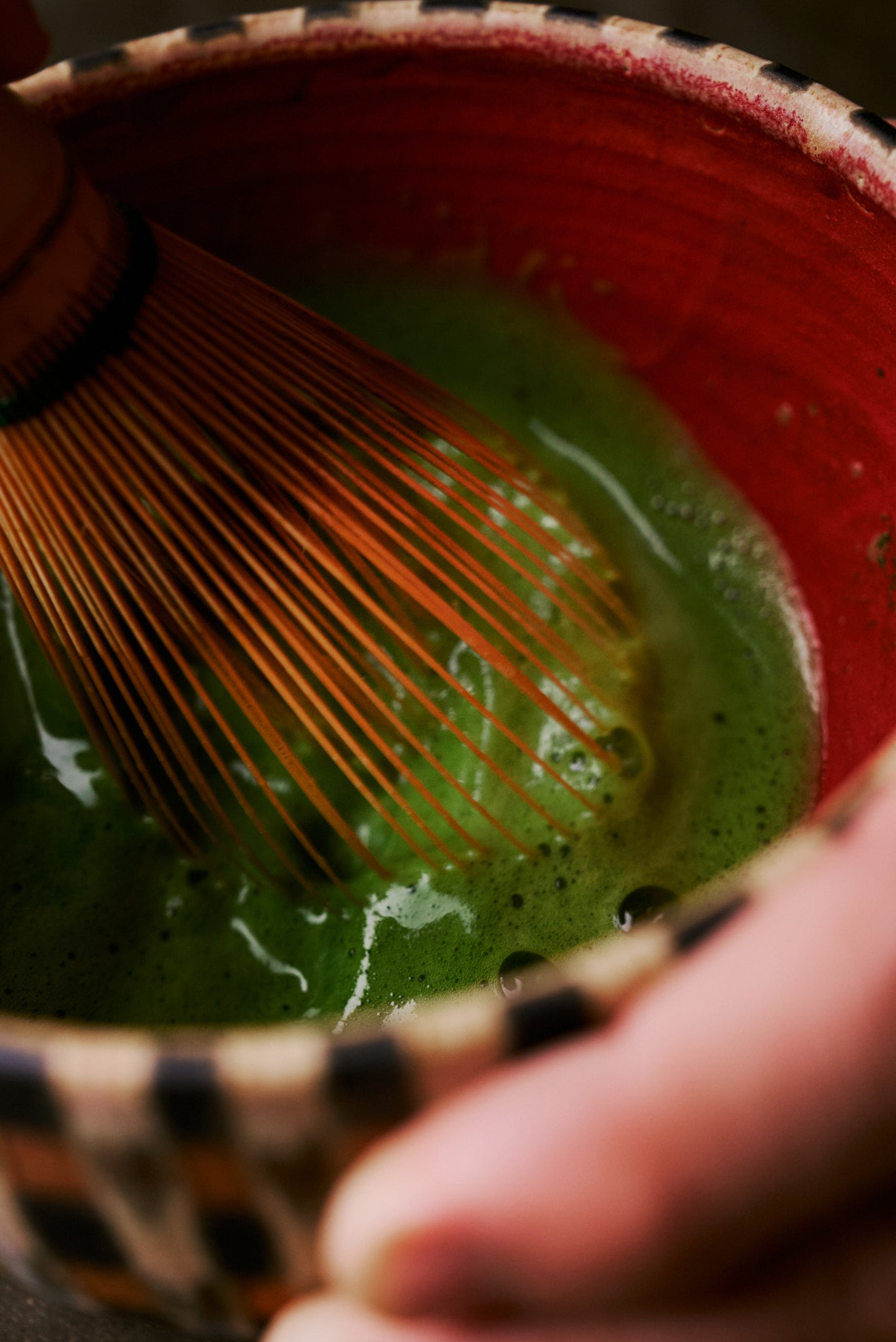 Whisking matcha with bamboo chasen in HIRAGOUSHI bowl - close-up of vibrant green tea and red glaze